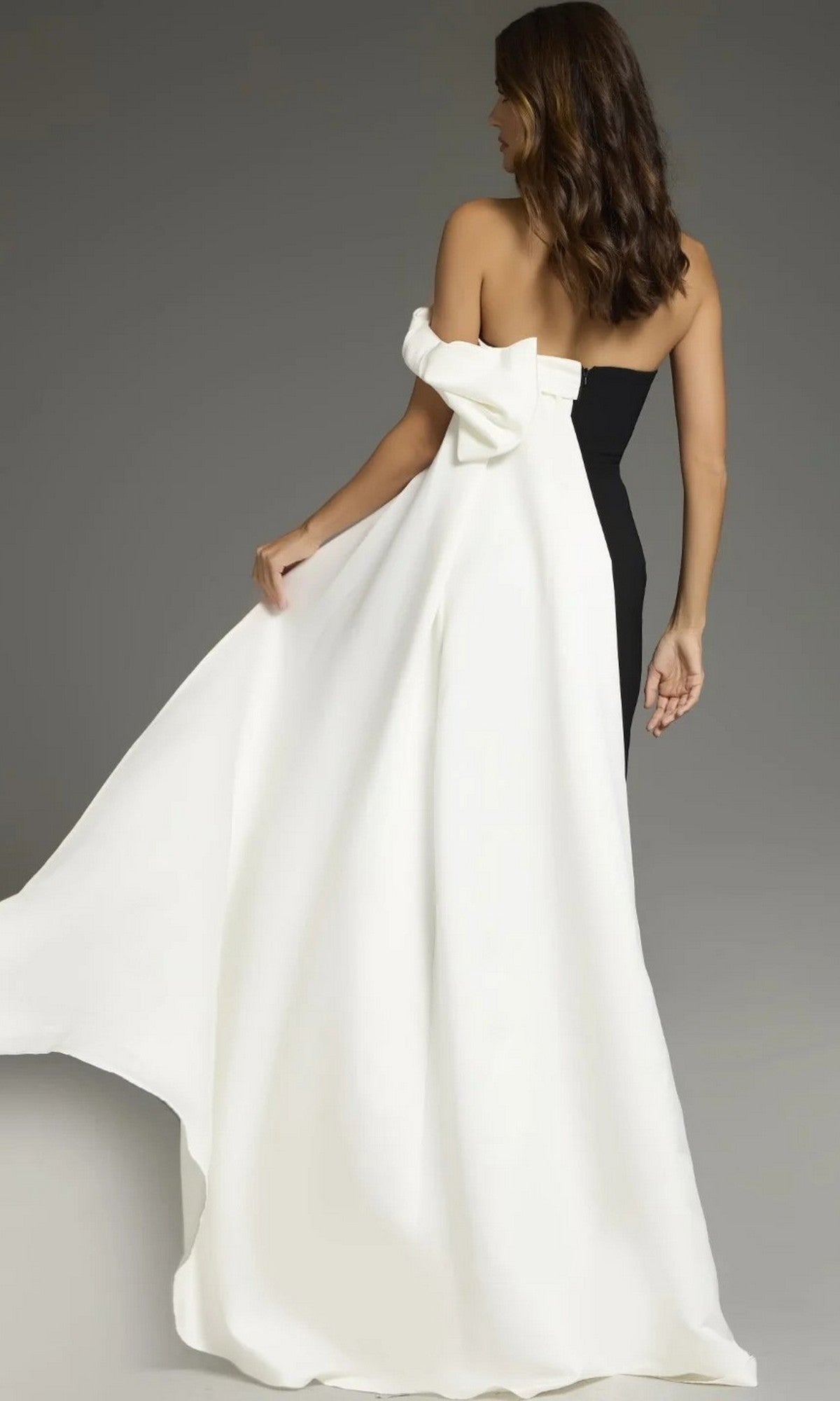 A woman stands against a plain background wearing the Jovani 38864 long formal dress, a strapless black and white gown featuring a large bow and a flowing white train.